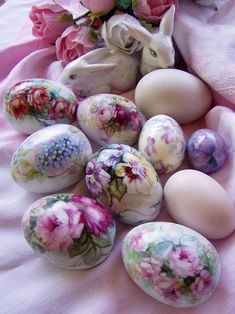 an assortment of painted eggs on a pink blanket with flowers in the middle and a white rabbit figurine next to it