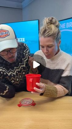 a man and woman sitting at a table looking at a red cup