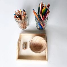 an assortment of colored pencils are in a glass jar and on a wooden tray