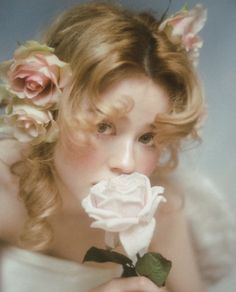 a woman with flowers in her hair is holding a rose up to her mouth and looking at the camera