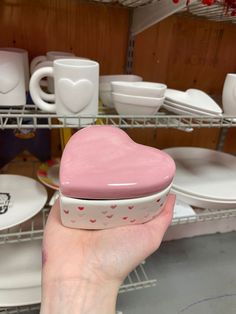 a hand holding a pink heart shaped box in front of white plates and mugs