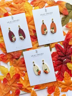 three pairs of earrings with flowers on them sitting next to autumn leaves and acorns