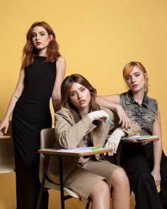 three young women sitting at desks in front of a yellow wall, one wearing a black dress