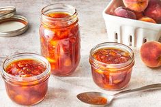 three jars filled with peach jam next to two spoons and a container full of peaches