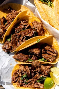 three tacos with shredded meat and garnishes on paper next to lime wedges