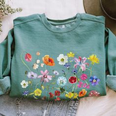 a green sweatshirt with colorful flowers on the front and side, sitting on top of jeans