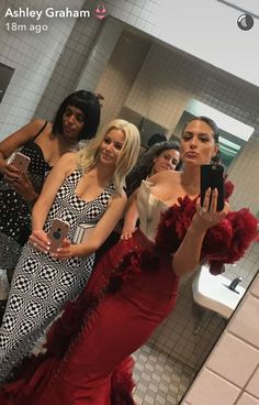 four women in dresses taking a selfie in a bathroom mirror with their cell phones