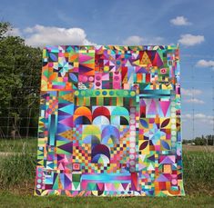 a colorful quilt is hanging on a wire fence in front of some grass and trees