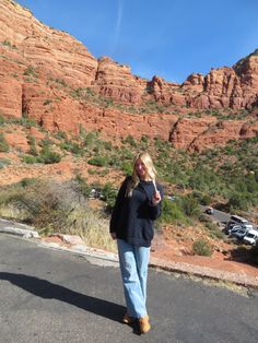 a woman standing on the side of a road talking on a cell phone with mountains in the background