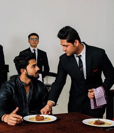 two men in suits and ties are eating at a table with another man looking on