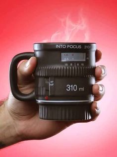 a hand holding a camera lens mug with steam rising out of it's side