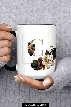 a woman holding a white coffee mug with flowers on it and the letter c in gold