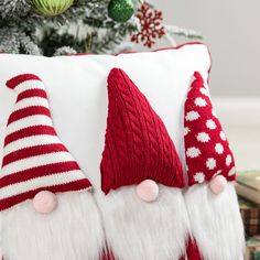 three red and white knitted christmas gnome hats sitting on top of a pillow in front of a christmas tree