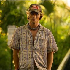 a man wearing a hat and shirt standing in front of some trees with his hands on his hips