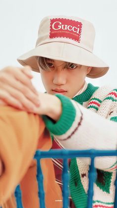 a young boy wearing a gucci hat and sweater, leaning against a blue fence