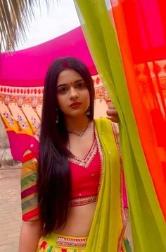 a woman wearing a yellow and green sari standing next to a colorful curtain in front of a palm tree