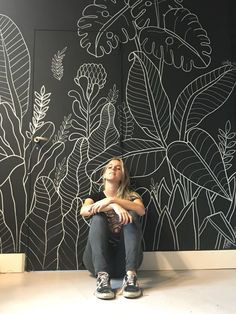 a woman sitting on the floor in front of a wall with plants drawn on it