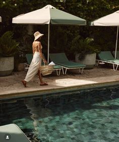 a woman in a white dress and hat walking by an outdoor pool with sun umbrellas