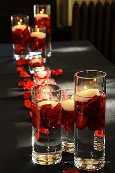 candles are lined up on a long table with red rose petals in glass vases