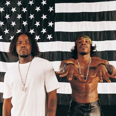 two men standing next to each other in front of an american flag with one pointing at the camera