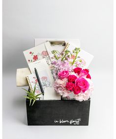 a black box filled with pink and white flowers next to a notepad, pen and pencil