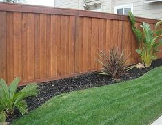 a wooden fence in front of a house with green grass and plants next to it