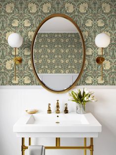 a white sink sitting under a mirror next to a gold faucet and wallpaper