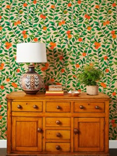 a wooden dresser with a lamp on top of it next to a floral wallpaper