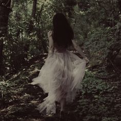 a woman in a white dress walking through the woods with her back to the camera