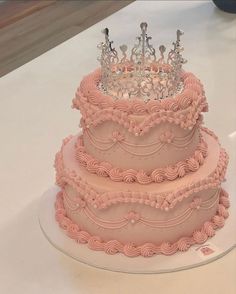 a three tiered cake with pink frosting and a crown on top is sitting on a table