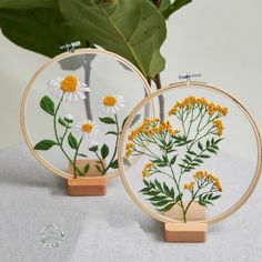 two cross stitch hoop paintings with yellow and white flowers in them on a table next to a potted plant