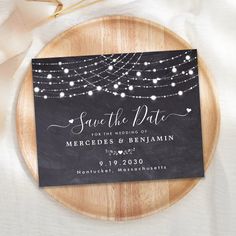 save the date card on a wooden plate with string lights and white ribbon around it