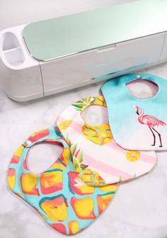 three bibs sitting next to a machine on a white counter with an ironing board in the background