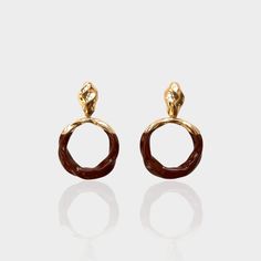 The Saddle Earrings present a unique blend of rustic charm and modern design. Each earring features a rich, saddle-brown circular drop with a marbled finish, giving the appearance of luxurious leather. The drop is framed by a glossy, twisted gold-tone setting that elegantly loops into the post. With their earthy tones and contemporary style, these earrings are versatile enough to enhance a casual daytime look or add an eclectic touch to an evening outfit. Whether for a day at the races or a nigh Luxury Brown Round Earrings, Nickel-free Brown Brass Earrings, Brown Patina Dangle Earrings, Luxury Gold-tone Brass Earrings, Artisan Brown Patina Earrings, Brown Earrings, Saddle Brown, Adventure Style, Evening Outfits