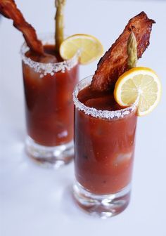 two cocktails with bacon and lemon garnish sit on a white table top