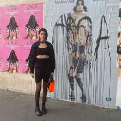 a woman standing in front of a wall with posters on it and holding an orange purse