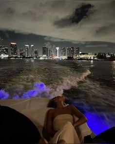 a woman laying on top of a boat in the water at night with city lights behind her