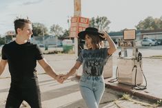 two people holding hands and walking down the street in front of an old gas station