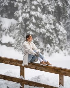 a woman sitting on top of a wooden rail in the snow next to a forest