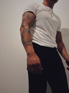 a man in white shirt and black pants with tattoos on his arm standing next to a wall