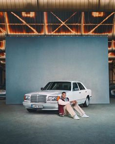 a man and woman sitting next to a white car in front of a large wall