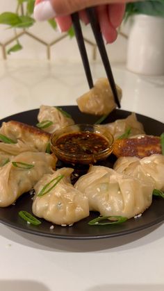 a person holding chopsticks over some dumplings on a black plate with dipping sauce