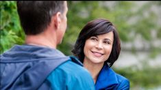 a man and woman standing next to each other smiling at each other with trees in the background