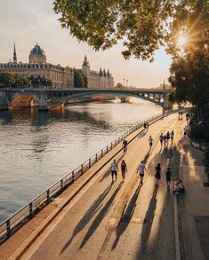 many people are walking on the sidewalk by the water and bridge in the city at sunset