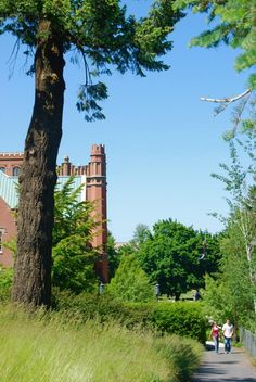 two people are walking down the path in front of an old brick building and tall trees