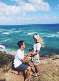 a man kneeling down next to a woman on top of a cliff near the ocean