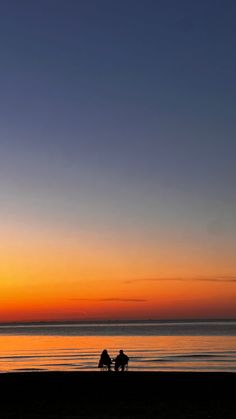 two people sitting on a bench at the beach watching the sun go down over the ocean