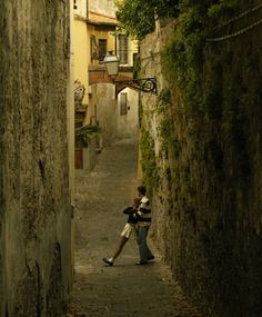 two people walking down an alley way with buildings on either side and trees growing up the wall