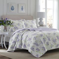 a white bed with purple flowers on it in a room next to a table and window