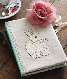 an embroidered book with a pink rose on top and a teacup next to it